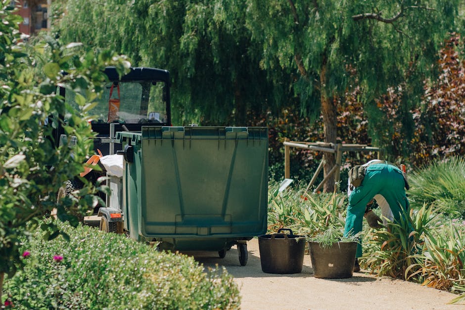 A person wearing a traditional conical hat and dark clothing is walking along a paved pathway in a park setting, pulling a green wheeled bin labeled 'Garden Waste' behind them. The area features lush 