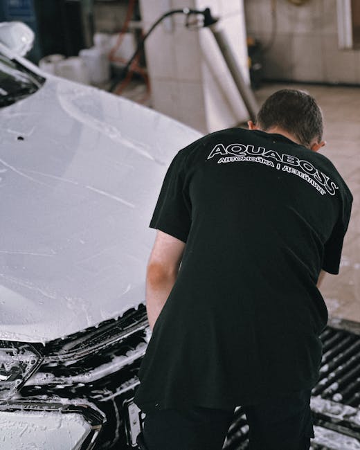 A person with short dark hair wearing a black T-shirt featuring the logo and text for Aquasox Automotive, is working on a silver car in a garage or workshop setting. The car appears to be undergoing m