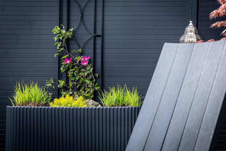 A modern outdoor garden scene featuring a rectangular, black, ribbed planter box filled with lush, bright green grass-like plants and small yellow flowering plants at the center. Behind the planter, a